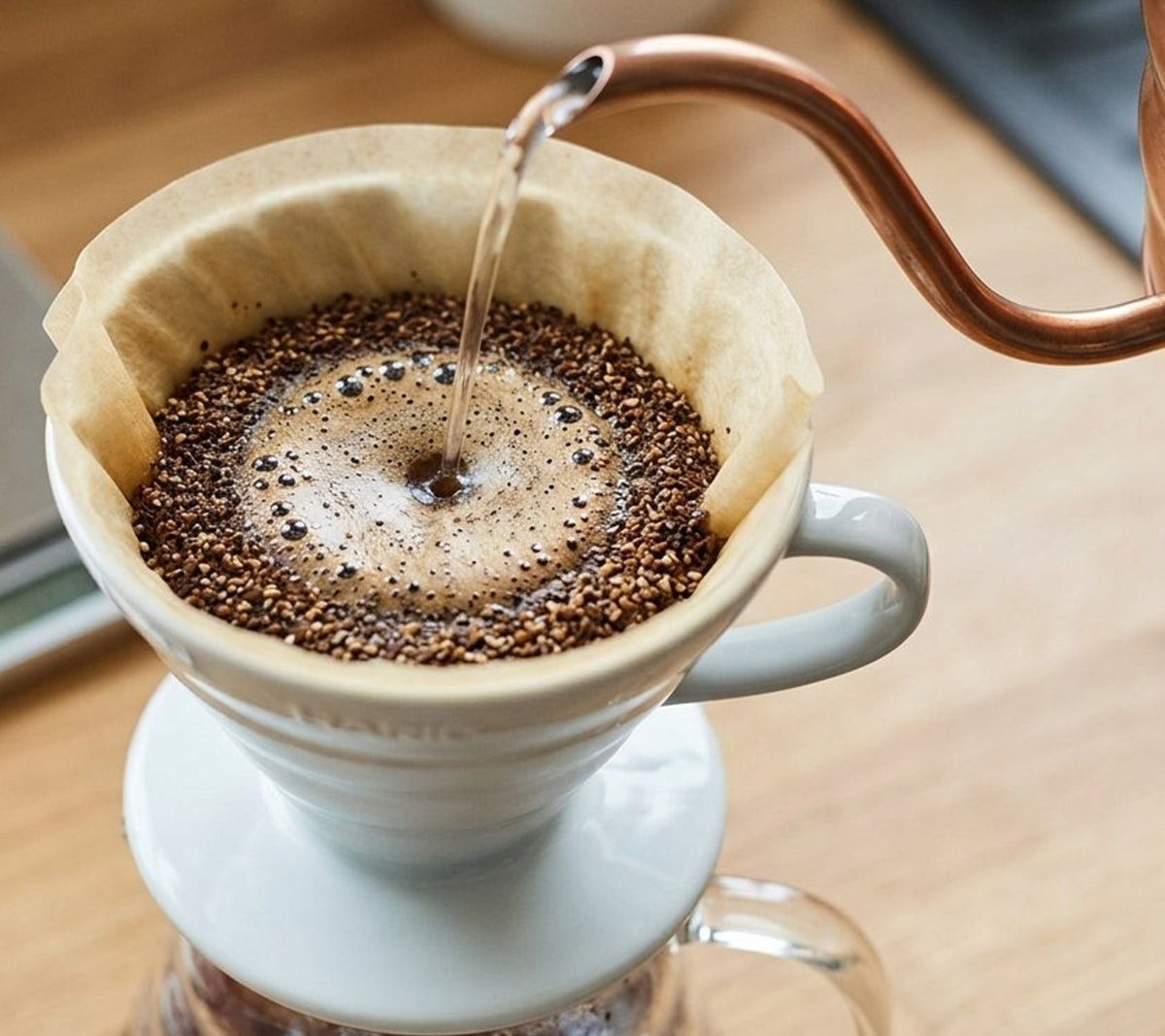 Pour-over coffee preparation with hot water being poured from a copper gooseneck kettle into a white ceramic dripper filled with coffee grounds and paper filter, set on a wooden surface.