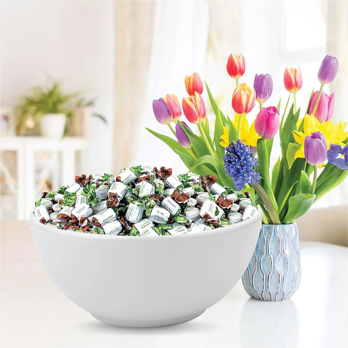 A white ceramic bowl filled with individually wrapped chocolates sits on a white table next to a blue patterned vase holding colorful tulips and a purple hyacinth in a bright, softly blurred indoor setting.