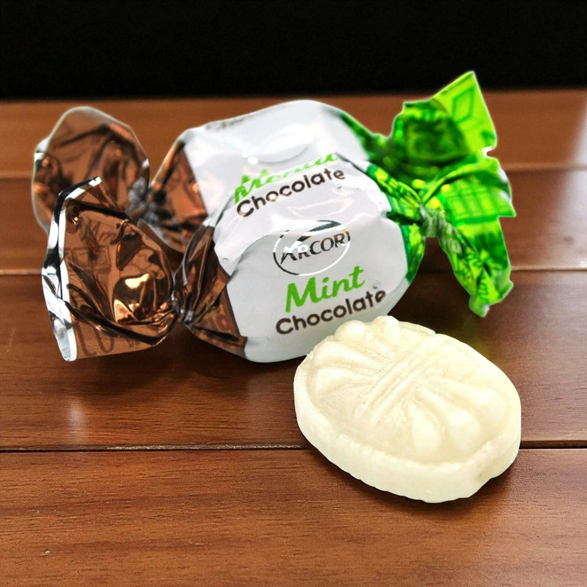 A wrapped mint chocolate candy lies on a wooden table with one unwrapped pale mint chocolate in front. The wrapper is white with green and brown foil sections and visible text including 'Mint Chocolate' and 'AnCOr'.